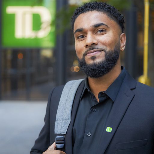 A student smiles while holding a laptop and phone outside on a college campus.