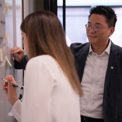A colleague writes on a whiteboard while brainstorming with another colleague.